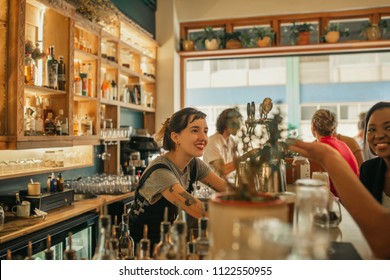 Smiling young female bartender standing behind the counter of a trendy bar taking drink orders from customers - Powered by Shutterstock