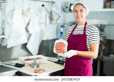 Smiling young female butcher demonstrating freshly formed ground beef hamburger patties in plastic tray, prepared for sale in meat processing room of local shop - Powered by Shutterstock