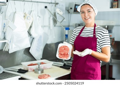 Smiling young female butcher demonstrating freshly formed ground beef hamburger patties in plastic tray, prepared for sale in meat processing room of local shop - Powered by Shutterstock