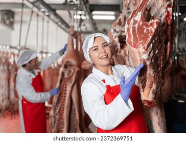 Smiling young female butcher shop worker checking slabs of fresh raw beef meat hanging on hook frame in cold storage room - Powered by Shutterstock