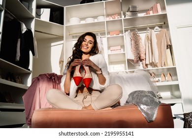 Smiling Young Female In Casual Clothes Looking At Camera While Sitting On Comfortable Couch And Showing New Red Shoes Against Shelves In Walk In Closet At Home