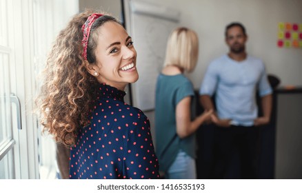 Smiling Young Female Designer Looking Over Her Shoulder During An Office Meeting With Colleagues Standing In The Background