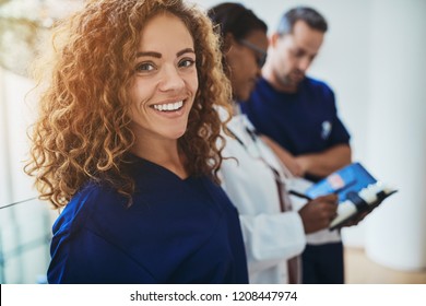 Smiling Young Female Doctor Standing In A Hospital Corridor With Two Medical Colleagues Talking In The Background