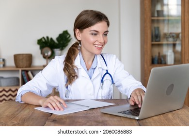 Smiling Young Female Doctor With Stethoscope Working On Laptop, Writing Report Or Email. Happy Millennial Woman GP Typing On Computer, Using Medical App, Consulting Patient Online, Filling Health Card