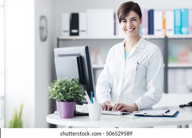 Smiling Young Female Doctor Working At The Clinic Reception, She Is Using A Computer And Writing Medical Reports