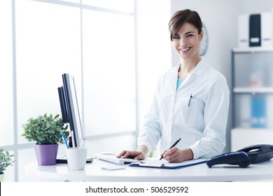 Smiling Young Female Doctor Working At The Clinic Reception, She Is Using A Computer And Writing Medical Reports