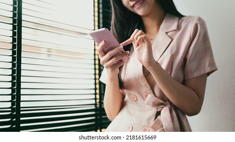 Smiling Young Female Employee Standing Near Office Window And Typing Text Message On Mobile Phone