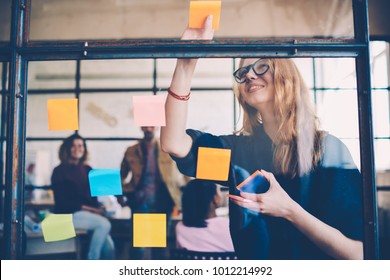 Smiling young female employee in stylish spectacles using stickers for creation strategy of working project during brainstorming session, skilled team member making notes presenting idea of startup - Powered by Shutterstock