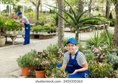 Smiling young female floriculturist in striped shirt and blue overalls happily displaying pink blooming Rhaphiolepis indica Springtime bushes in sunlit open garden center - Powered by Shutterstock