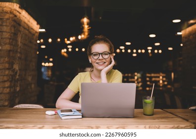 Smiling young female freelancer with TWS earbuds and eyeglasses look at camera with hand at chin while listening to music during break at table with laptop smartphone diary pen glass of juice in cafe