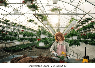 Smiling young female ginger florist standing at greenhouse with trowel in hands and planting blooming flower into flowerpot. Happy redhead horticulturist replanting and cultivating flowers at hothouse - Powered by Shutterstock