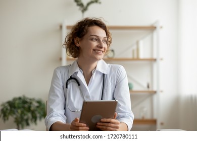 Smiling young female professional doctor holding digital tablet looking away sitting at workplace desk. Happy beautiful woman physician wearing white coat and stethoscope dreaming in medical office.
