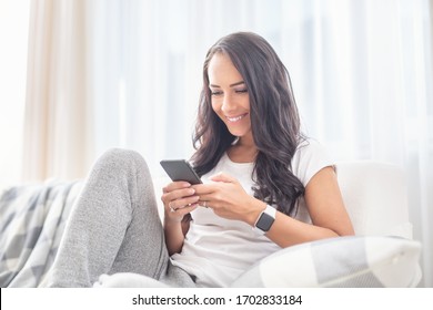 Smiling young female sitting on a couch in a living room smiling into the phone spending time online during a lockdown. 