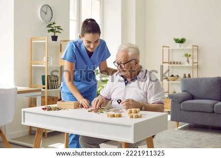 Smiling young female social worker and old man playing with jigsaw puzzle in nursing home. Caregiver in medical uniform helps elderly man to put together puzzles and letters. Old age concept.