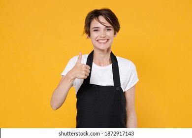 Smiling Young Female Woman 20s Barista Bartender Barman Employee In White T-shirt Apron Work In Coffee Shop Posing Showing Thumb Up Looking Camera Isolated On Yellow Color Background Studio Portrait