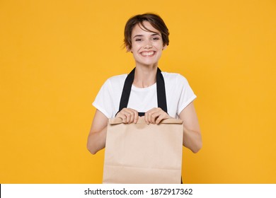 Smiling Young Female Woman 20s Barista Bartender Barman Employee In Apron Hold Brown Empty Blank Craft Paper Bag For Takeaway Mock Up Looking Camera Isolated On Yellow Wall Background Studio Portrait