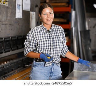 Smiling young female worker in casual plaid shirt, jeans and gloves posing at workplace in metalworking shop, surrounded by tools and machines, holding tape measure, ready to mark workpieces.. - Powered by Shutterstock