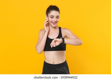 Smiling young fitness sporty woman 20s wearing black sportswear posing working out training wearing smart watch on hand listen music with air pods isolated on yellow color background studio portrait