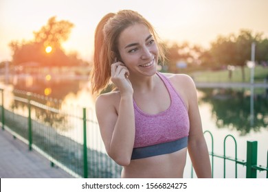 Smiling Young Fitness Woman With Airpods Listening To Music During Her Outdoor Workout In The Park