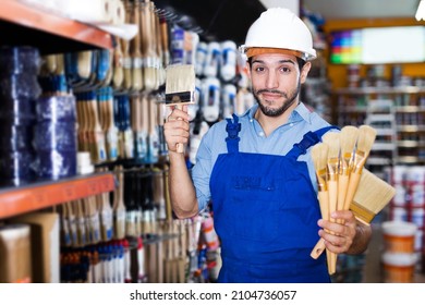 Smiling Young Foreman In Blue Overalls Choosing Brushes In Paint Store