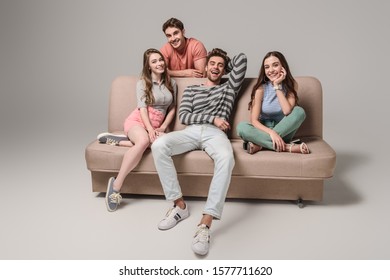 Smiling Young Friends Sitting On Sofa On Grey 