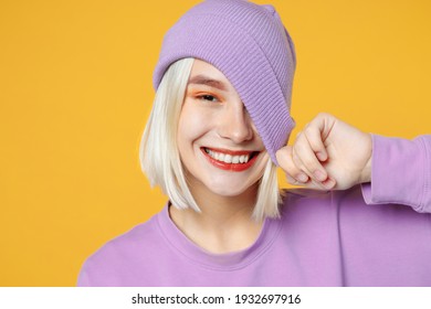 Smiling young funny blonde caucasian woman 20s bob haircut bright makeup wearing basic casual purple shirt covering eye with violet beanie hat isolated on yellow color background studio portrait