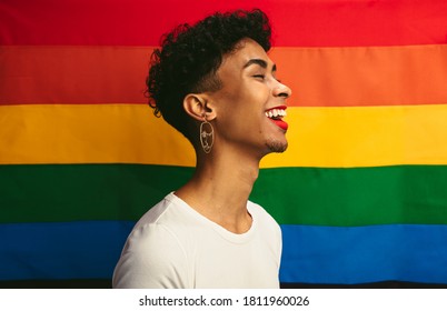 Smiling young gay man with make up standing against pride flag. Man with red lip stick and earring laughing in front of rainbow flag of gay pride.