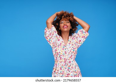 Smiling young girl with afro hairstyle and glamour makeup posing in floral dress on blue studio background, looking at the camera. A lot of copy space.