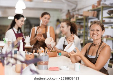 Smiling young girl in apron painting on handmade ceramic bowl with brush in pottery workshop - Powered by Shutterstock