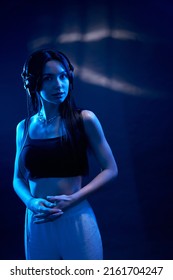 Smiling Young Girl In Black Top Listening To Music While Mysteriously Looking At Camera Indoor. Cute Female Dj Wearing Headphones Standing In Dark Blue Neon Light In Studio. Concept Of Lifestyle. 