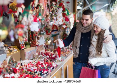 Smiling young girl with boyfriend choosing Christmas decoration at fair. Focus on man