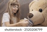 Smiling young girl with long blonde hair pretends to give tea to a large teddy bear. She holds a cup in a charming imaginary play scene.
