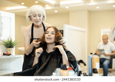 Smiling young girl with long brown hair sitting in chair in hairdressing salon, discussing with experienced professional female hairstylist while choosing new hairdo - Powered by Shutterstock