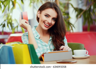Smiling young girl with long brown hair wearing casual shirt sitting in cafe with colorful shopping bags, cup of coffee and tablet, holding credit card, online shopping concept, portrait.
