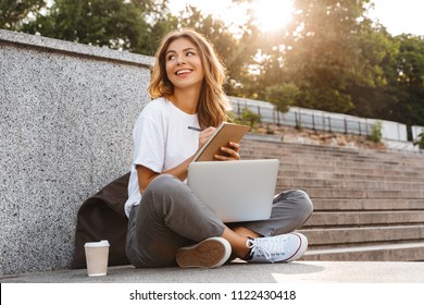 Smiling young girl making notes in notepad while sitting outdoors with laptop computer and coffee cup - Powered by Shutterstock