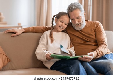 A smiling young girl sits closely to her grandfather as they share a moment of storytelling. The grandfather is focused on the book in the girls hands, both seem wrapped up in the tale - Powered by Shutterstock