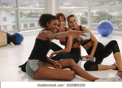 Smiling Young Girls Taking Selfie In Gym. Three Female Friends In Healthclub Taking Selfie With Smart Phone.