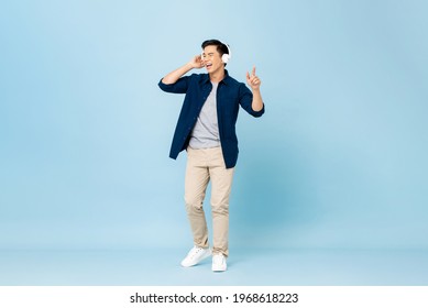 Smiling young handsome Asian man listening to streaming music on wireless headphones in isolated light blue studio background