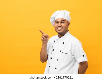 Sonriente Joven apuesto chef asiático con uniforme y sombrero apuntando con el dedo sobre el fondo amarillo espacio en blanco para el texto. Hombre cocinero o panadero El cocinero de la cocina