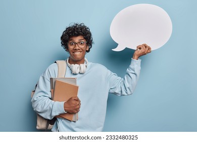 Smiling young happy Hindu male wearing casual hoodie with leather bag and headphones standing isolated on blue background holding books and white speech bubble for your promotion.Education concept - Powered by Shutterstock