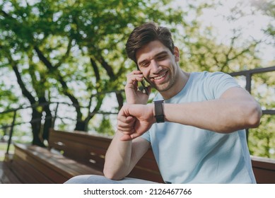 Smiling young happy man wear blue t-shirt sit on bench talk speak on mobile cell phone check time on smart watch rest relax in sunshine spring green city park outdoor on nature Urban leisure concept.