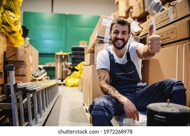 Smiling Young Hardworking Tattooed Bearded Blue Collar Worker In Overalls Sitting In Storage Surrounded By Boxes And Giving Thumbs Up. Import And Export Firm Interior.