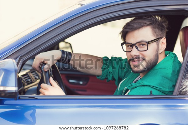 Smiling young hipster man buyer sitting\
in his new car excited ready for a trip on a city street\
background. Personal transportation auto purchase\
concept