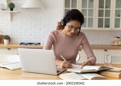 Smiling Young Hispanic Caucasian Woman In Glasses Wearing Headphones With Mic Listening Online Educational Lecture Or Holding Video Call With Teacher, Writing Notes, Distant E-learning Concept.