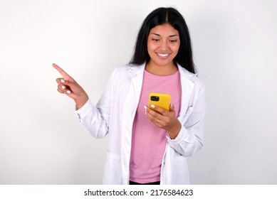 Smiling Young Hispanic Doctor Girl Wearing Coat Over White Background Pointing Finger At Blank Space Holding Phone In One Hand