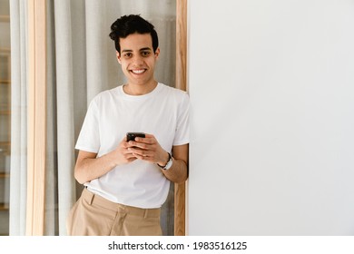 Smiling Young Hispanic Man Standing Leaning On A Wall At Home, Holding Mobile Phone