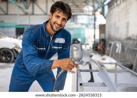 A smiling young Hispanic mechanic points at a spray gun nozzle beside a car bumper in a repair shop.