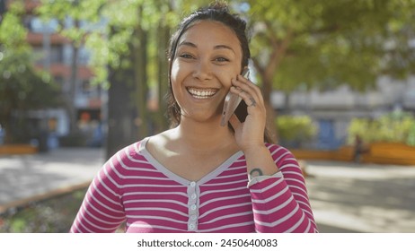 A smiling young hispanic woman chats on a phone in a sunlit park, exuding joy and beauty amidst greenery and urban surroundings. - Powered by Shutterstock