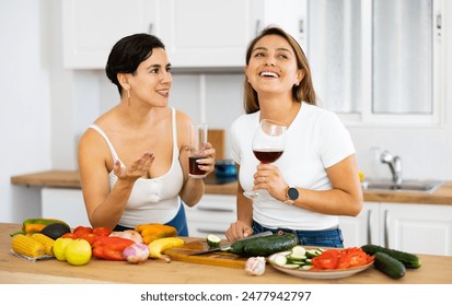 Smiling young Hispanic woman cooking with sister in home kitchen, chatting cheerfully and drinking wine while preparing vegetable salad. Happy family relationship concept.. - Powered by Shutterstock