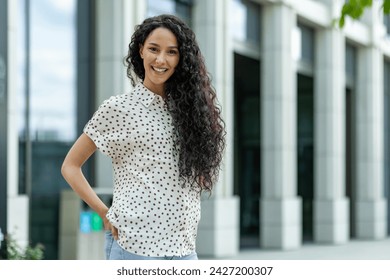 Smiling young Hispanic woman with curly hair enjoying her lunch break outside an office. Confident and professional. - Powered by Shutterstock